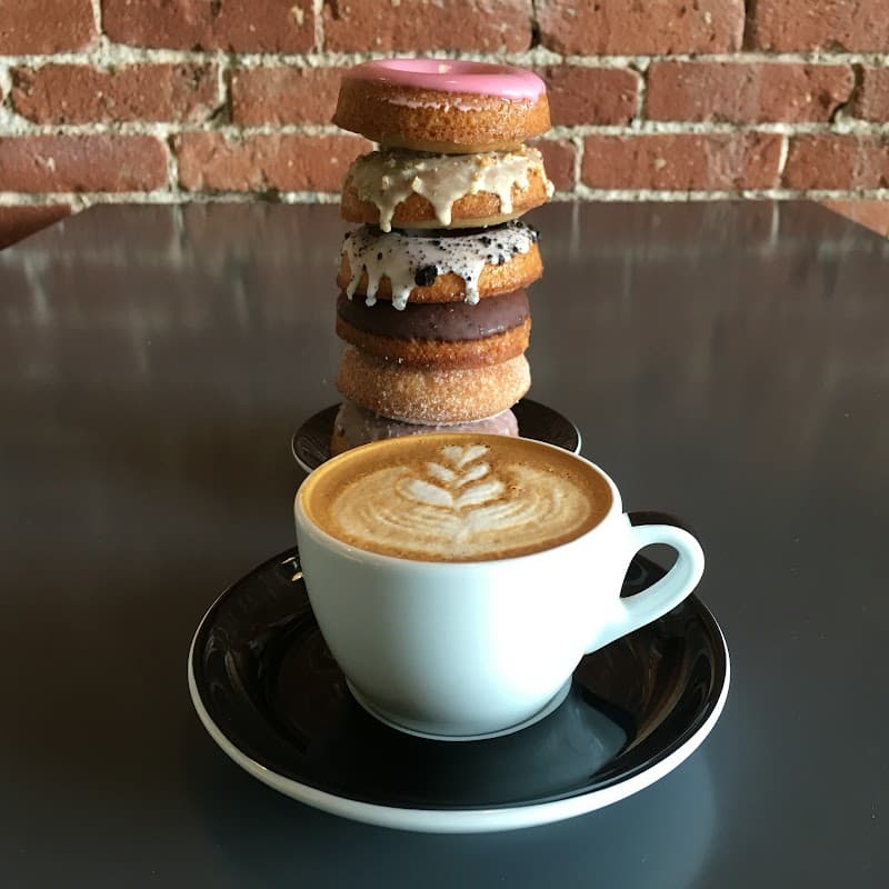 Latte with leaf art beside stacked assorted donuts on dark table against exposed brick wall 1